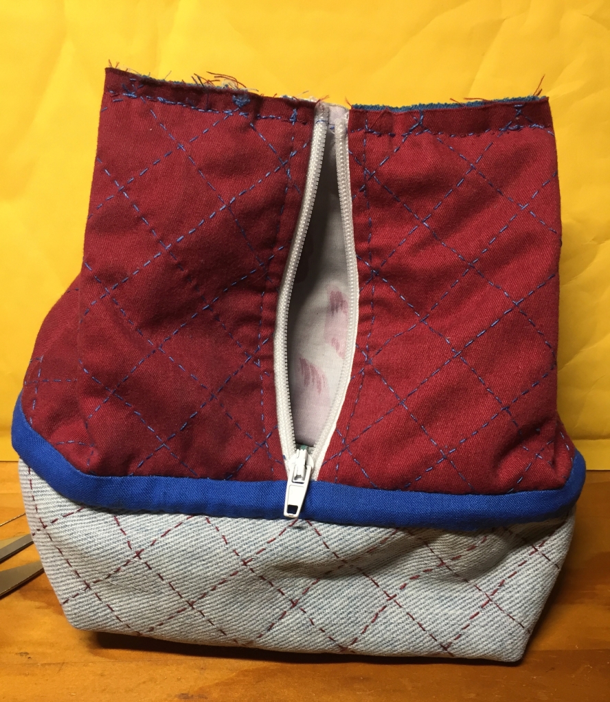 The unfinished pouch, with the folded-inwards top seam sewn, and the zipper open so the pastel chintz lining is visible. The joint with the bottom denim box-like shape is covered with a strip of blue fabric in the same shade as the quilting thread on the top half.