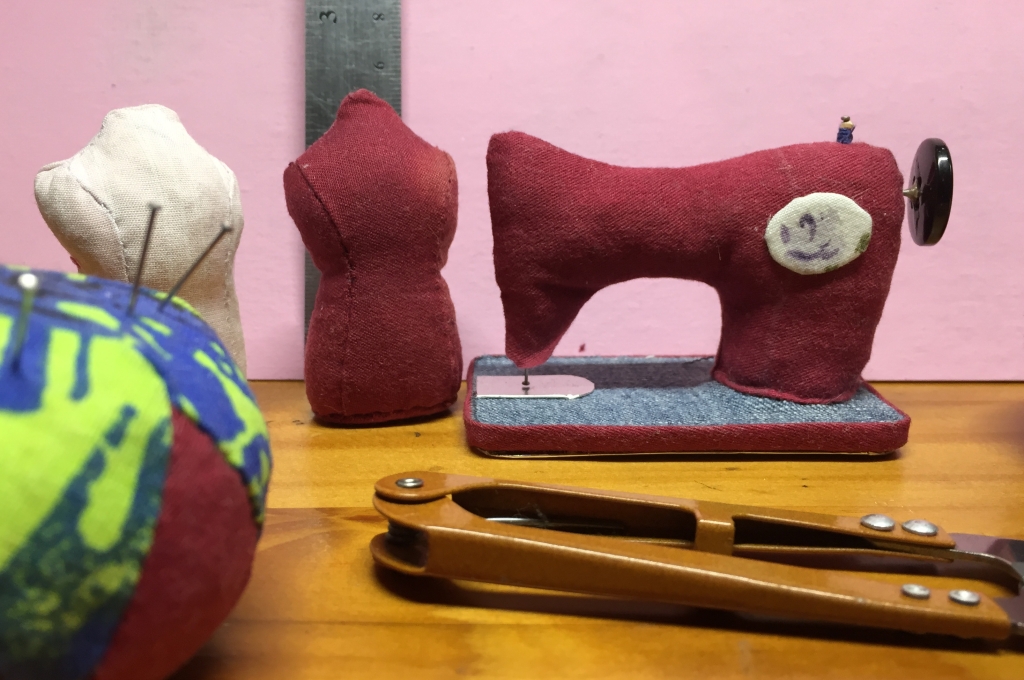 Close photograph of two seamstress dressforms about 2.5 inches tall, and a miniature old-fashioned "sewing machine" about 2 inches high by 4 inches long, all made of cotton fabric and stuffed tight. There's a button mounted on a pin to represent the wheel, and a tiny wooden thread spool, with thread on it, at the top.