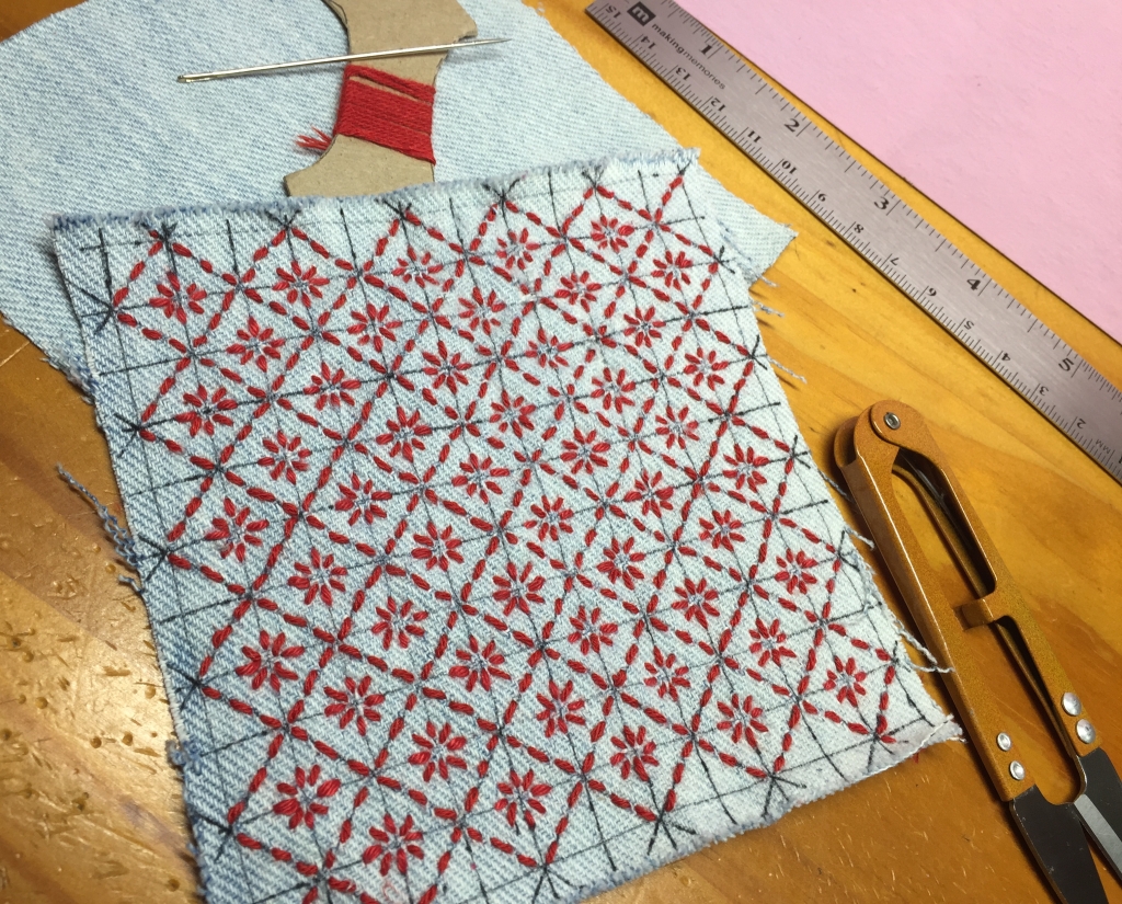 a square of denim with lines drawn in a grid, partially embroidered with a sashiko pattern in red thread; also visible, the red sashiko thread, the long sashiko needle, thread snips and a 6in ruler