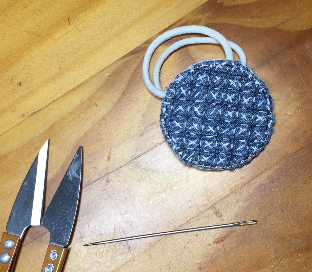 A circle of denim fabric showing a grid of white stitches; inside each square, there's a cross of stitches. Around the border there are a couple of rows of buttonhole stitches, which are holding the various layers of fabric together. Sewn to the back are two loops of elastic, smaller than the circle itself