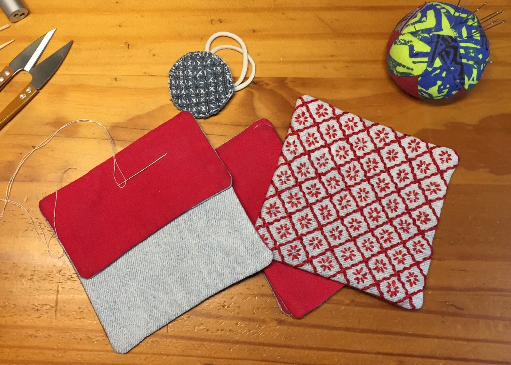 Starting to attach the finished pieces of the square denim pouch, some of the panels are on the desk, the red cotton lining visible, as is the sashiko style embroidered front panel with the red design on denim