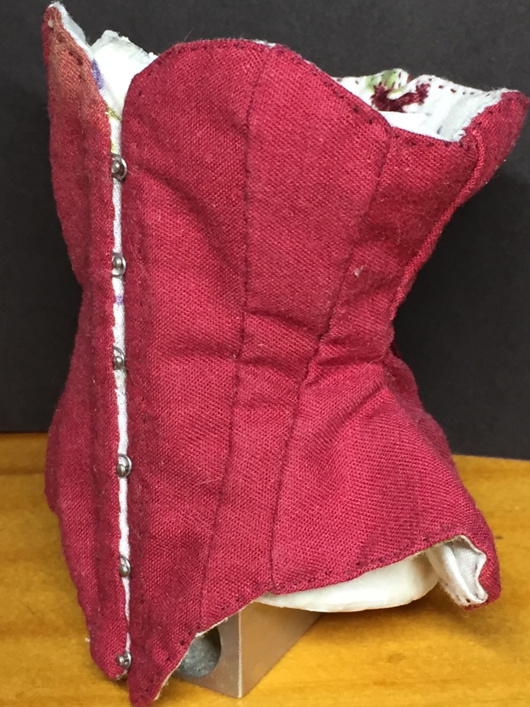 Three quarter profile view, showing the metal wire loops going around the pins on the other half of the busk. In the back, the lining shows the tiny topstitching in burgundy thread, as well as the top eyeletes in the back, with the laces.