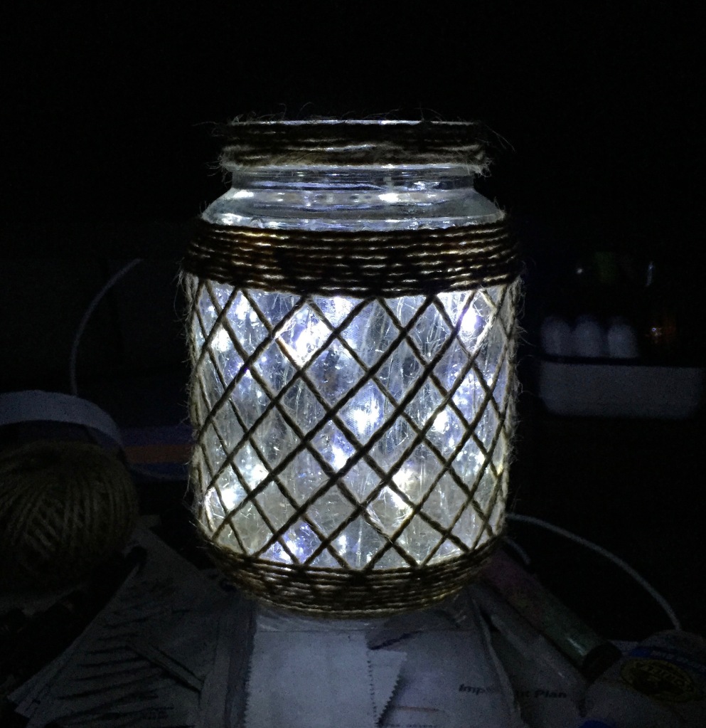 A glass jar covered in a lattice of jute twine, shot in the dark, with many small lights shining through the glass. The dark background is the untidy surface of my desk, with a ball of the twine clearly visible on the side.