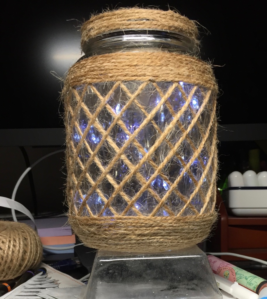A glass jar covered with a lattice of jute twine, with the fairly lights inside turned on; the thread at the mouth of the jar is now covered with jute twine glued around it.